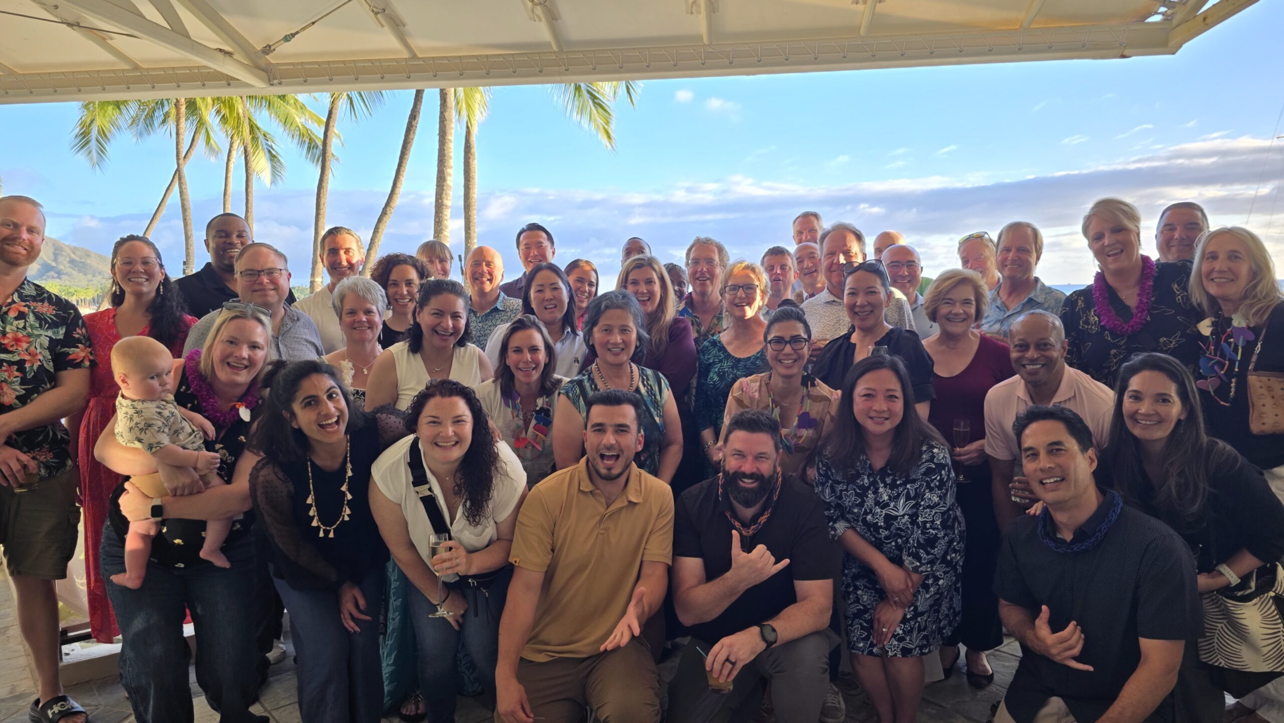 A large, diverse group of people gathered outdoors for a team photo under a canopy with palm trees and a bright sky in the background.