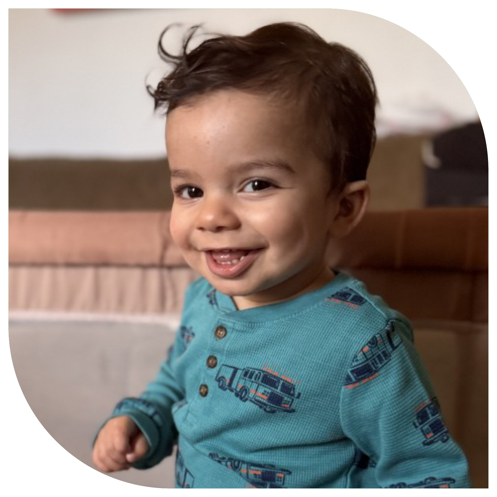 A toddler with dark hair and a teal shirt featuring vehicle prints smiles while standing in a cozy room. The atmosphere is warm and cheerful.