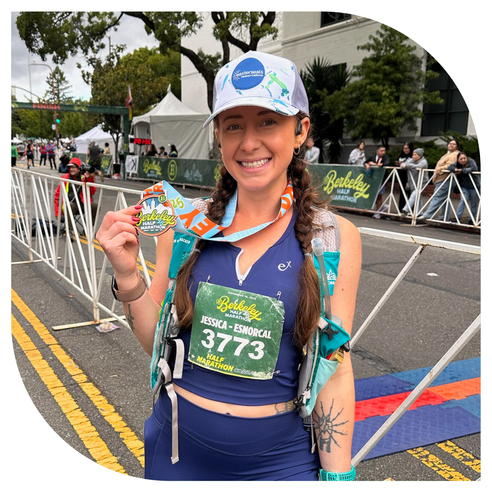 A woman smiles, holding a medal at a finish line. She wears a race bib with the number 3773, a cap, and athletic gear. The mood is triumphant and celebratory.
