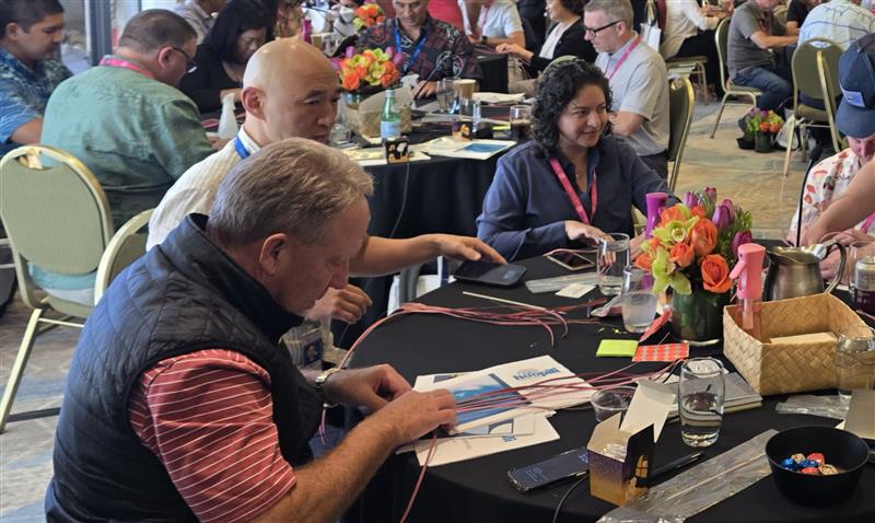 Two men sitting at a round table during a workshop, focused on a collaborative hands-on activity with papers and red string.