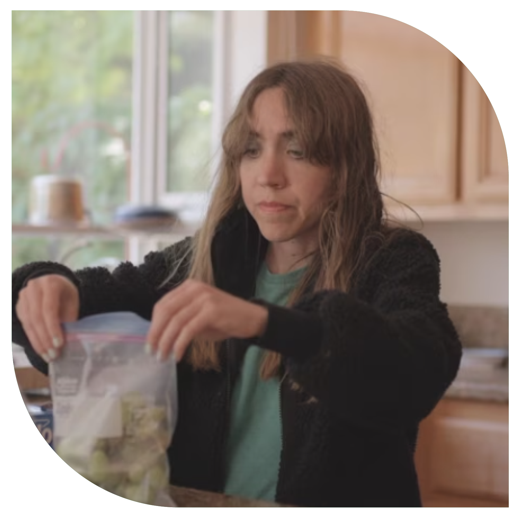 Woman with long hair prepares food in a bright kitchen, placing items in a clear bag. She wears a dark jacket and appears focused. Light and calm ambiance.