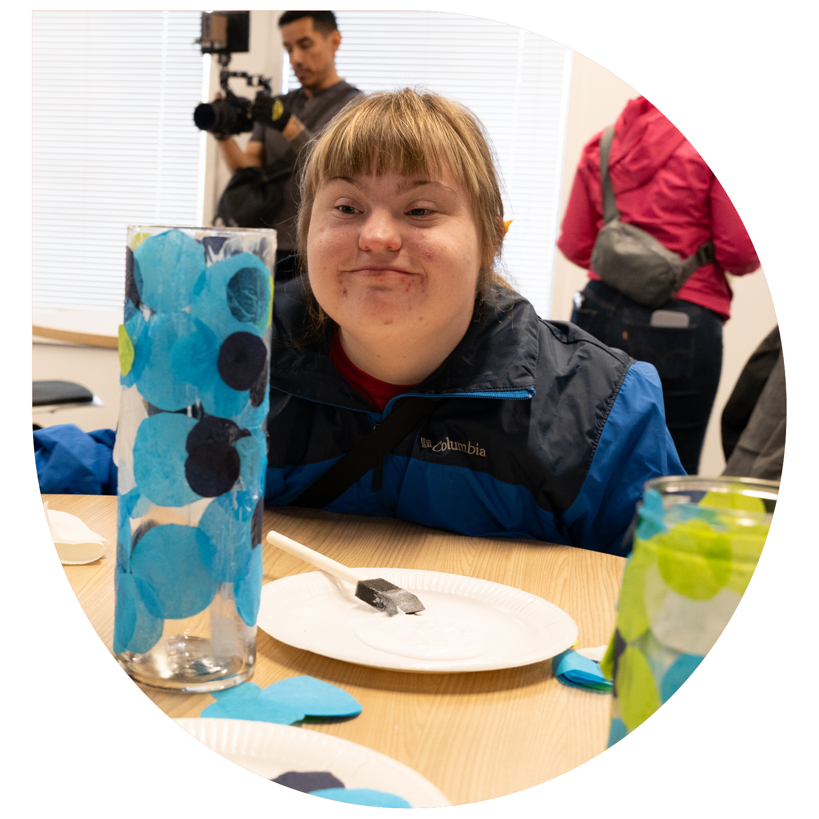 A person smiling while seated at a table with artistic materials, including a decorated vase with blue circles and paper plates. The atmosphere is creative and joyful.