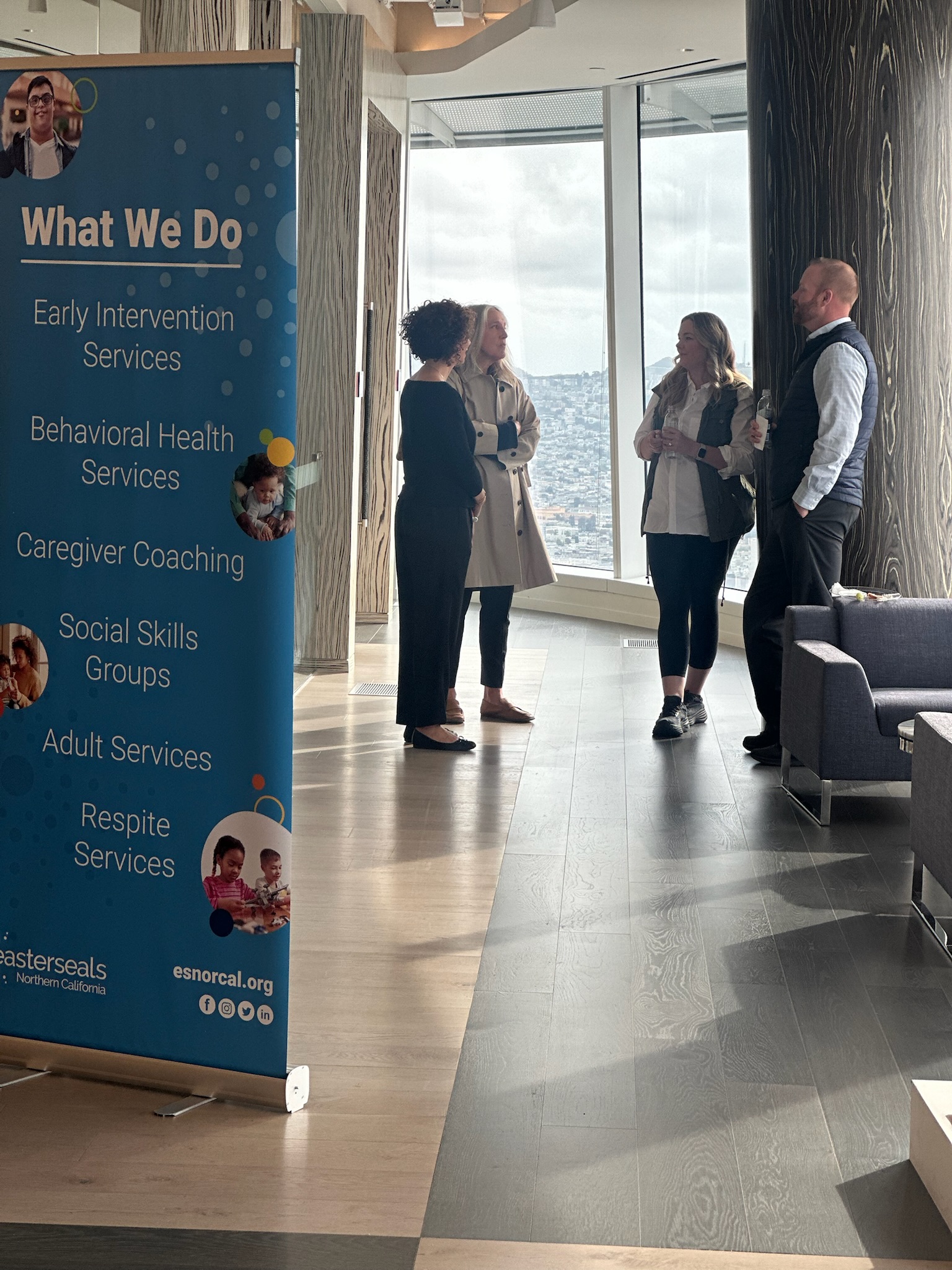 A candid shot of four professionals engaged in conversation near a large window overlooking a city, next to a blue banner detailing 
