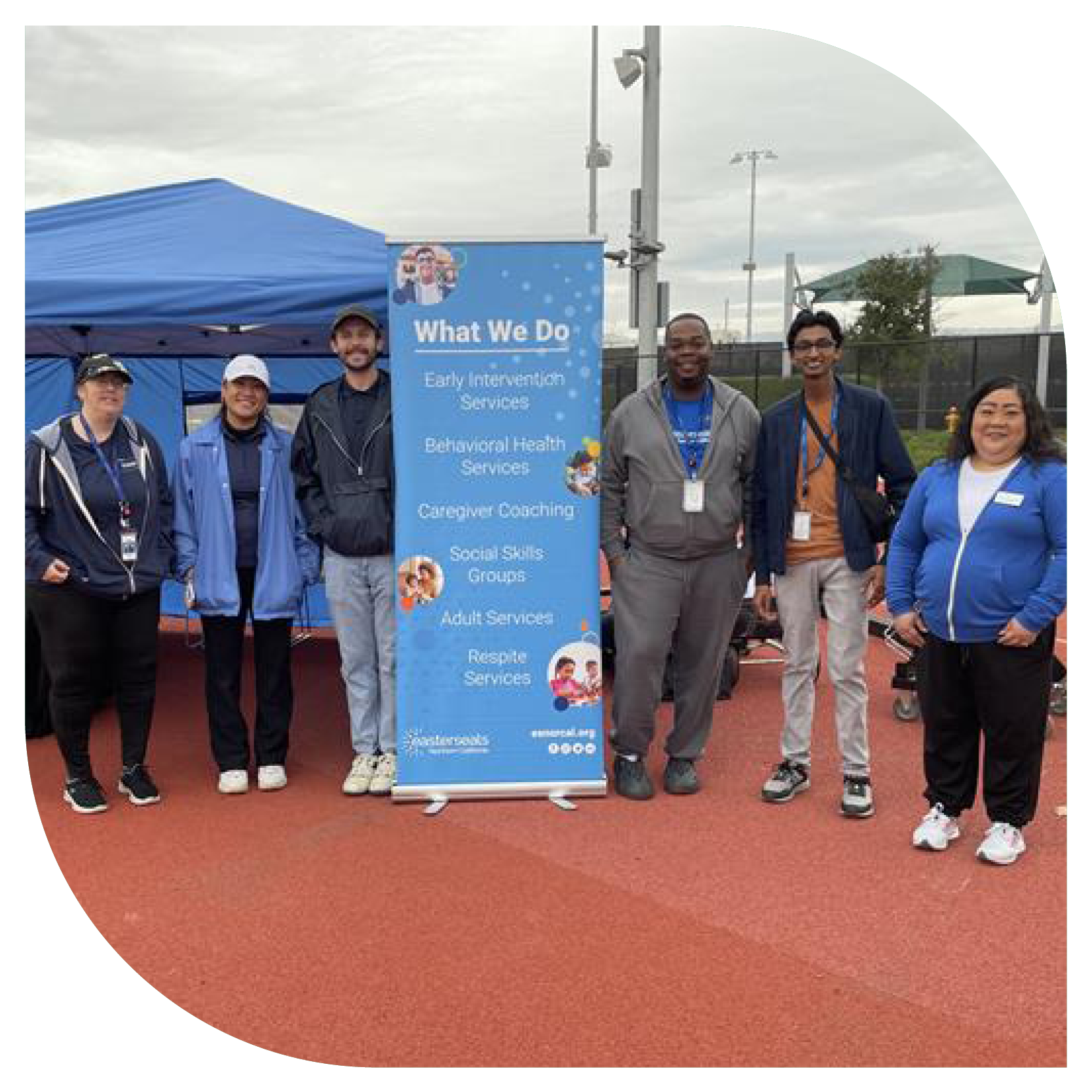 A group of six people stand on a sports track, smiling, in front of a blue information banner and tent. The sky is overcast, creating a casual, friendly atmosphere.
