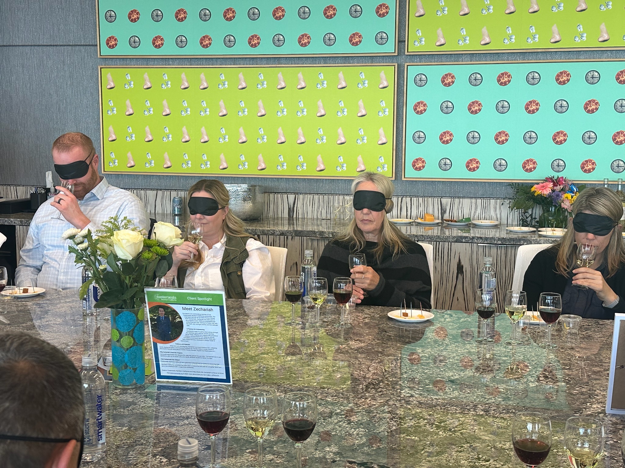 A group of people sitting at a long table wearing black blindfolds, participating in a guided sensory or wine-tasting experience.