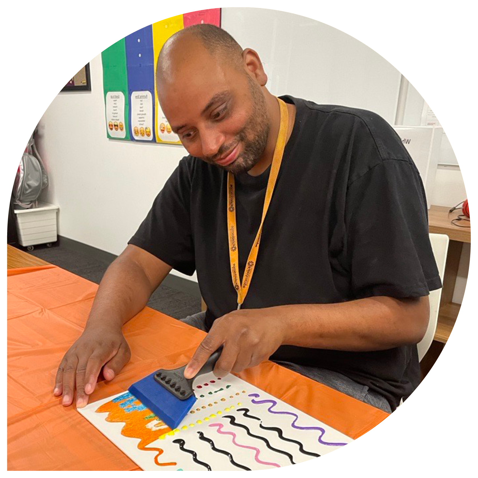 A man wearing a lanyard uses a blue brush to paint colorful wavy lines on paper. He's seated at an orange table, engaged and focused on the activity.